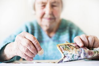 Eine Seniorin sitzt an einem Tisch, zählt Münzen in ihr Portemonaine. Ihr Gesichtsausdruck ist ernst.