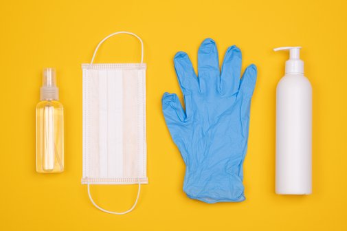 Eine Flasche mit Desinfektionsmittel, ein Mundschutz, ein blauer Gummihandschuh uind eine Flasche mit einer Lotion nebeneinander vor gelbem Hintergrund
