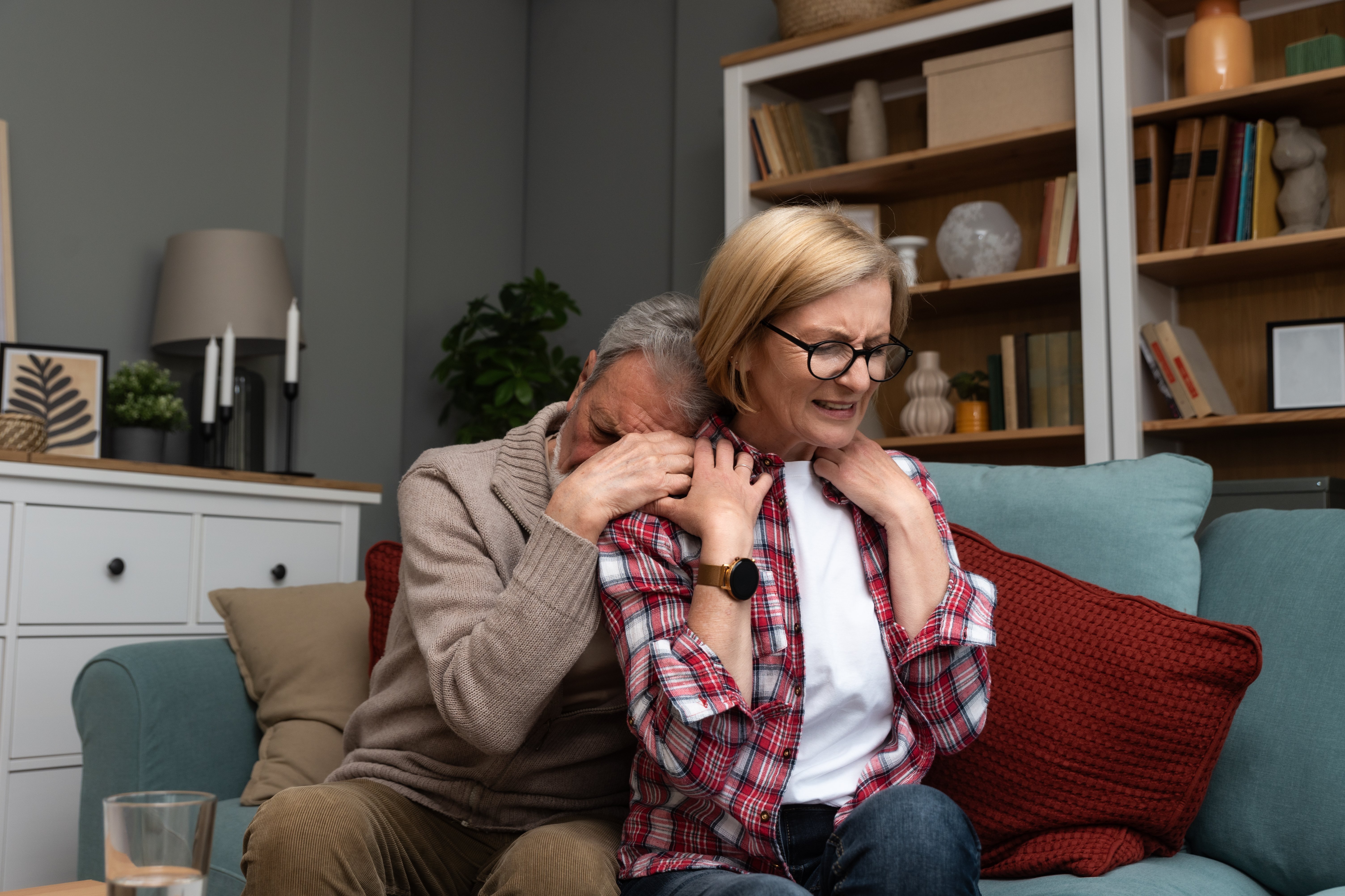 Ein &auml;lterer Mann umarmt eine &auml;ltere Frau von hinten, sie sitzen auf einem Sofa. Er hat sein Gesicht von hinten in ihrer Schulter vergraben. Sie h&auml;lt seine H&auml;nde, blickt geradeaus mit einem angestrengten, ernsten Ausdruck. Symbolfoto f&uuml;r Ersch&ouml;pfung in der Angeh&ouml;rigenpflege. 