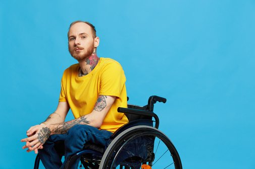 Ein junger Mann im gelben T-Shirt, der im Rollstuhl sitzt, vor blauem Hintergrund. Er blickt ernst in die Kamera.