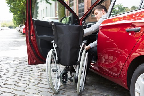 Ein junger Mann steigt von seinem Rollstuhl in sein Auto ein und setzt sich auf den Fahrersitz.