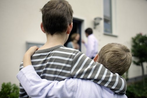 Zwei Jungen stehen Arm in Arm nebeneinander, im Hintergrund sieht man die Eltern streiten