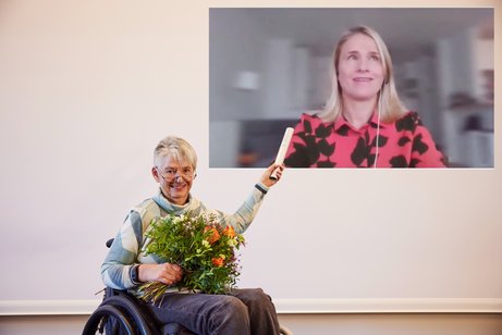 DBR-Staffelstab-Übergabe Dr. Sigrid Arnade hält den DBR-Staffelstab in die Höhe, wo auf einer Leinwand Verena Bentele digital zugeschaltet ist, und übergibt ihr damit symbolisch den DBR-Vorsitz für das nächste Jahr.