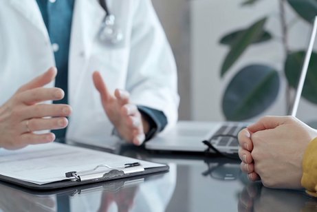 Bildausschnitt aus einem Arzt-Patientinnen-Gespr&auml;ch: Ein Arzt im wei&szlig;en Kittel mit einem Stethoskop um den Hals gestikuliet. Gegen&uuml;ber sitzt eine Patientin, hat die H&auml;nde auf dem Tisch zusammengelegt.