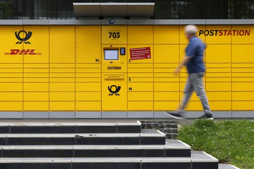 Eine Poststation von DHL. Ein Mann geht gerade daran vorbei. Im Vordergrund sieht man eine Treppe.