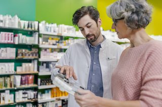 Ein Mitarbeiter einer Apotheke im weißen Arbeitskittel spricht mit einer älteren Frau über ein Medikament, dessen Packung er in der Hand hält.