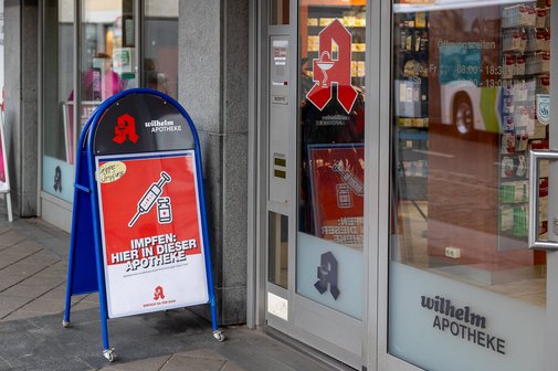 Eine Apotheke mit einem Aufsteller vor dem Eingang, der auf die M&ouml;glichkeit einer Grippeschutzimpfung hinweist. Das Schild tr&auml;gt die Aufschrift Impfen: Hier in dieser Apotheke und zeigt ein Symbol einer Spritze und eines Impfstoff-Fl&auml;schchens.
