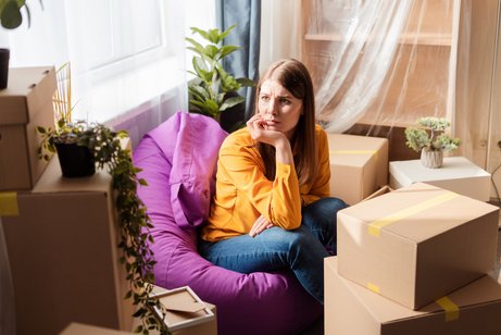 Haushalt auflösen Eine junge Frau sitzt auf einem Sitzsack in einer Wohnung zwischen Umzugskisten, sie blickt traurig und besorgt, hat das Kinn in die Hand gestützt.