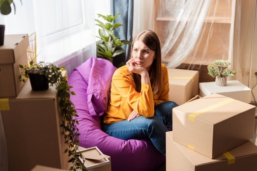 Eine junge Frau sitzt auf einem Sitzsack in einer Wohnung zwischen Umzugskisten, sie blickt traurig und besorgt, hat das Kinn in die Hand gestützt.