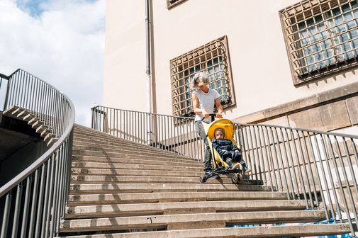 Frau mit Kinderwagen bei Hitze Eine Frau fährt mühevoll mit einem Buggy mit Kleinkind treppab, die Sonne scheint, sie wirkt erhitzt und angestrengt