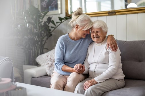 Pflegende Angehörige Eine ältere Frau sitzt mit einer noch älteren Seniorin auf einem Sofa, sie nimmt die Ältere liebevoll in dem Arm. Das Bild strahlt Nähe und Liebe aus.
