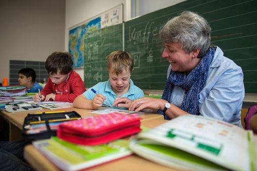 Blick in eine Schulklasse, eine Inklusionshelferin k&uuml;mmert sich um einen Jungen mit Trisomie 21. 
