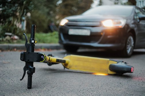 Ein gelber E-Scooter liegt umgest&uuml;rtzt auf der Stra&szlig;e. Ein Auto, dessen Scheinwerfer an sind, steht weniger Meter entfernt. 