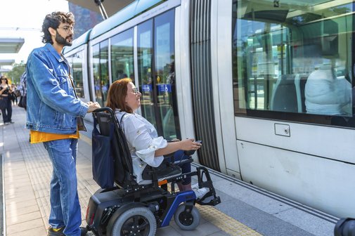 Junger Mann, der einer erwachsenen Frau im Rollstuhl beim Einsteigen in eine moderne Stra&szlig;enbahn an einer belebten Haltestelle hilft, um Barrierefreiheit und Inklusion zu unterstreichen