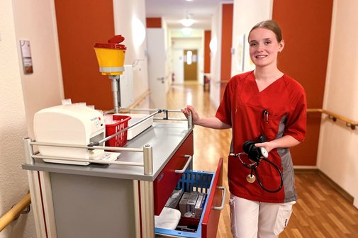 Das Foto zeigt Michelle. Sie steht neben einem Medikamentenwagen im Flur des Pflegeheimes und hält ein Blutdruckmessgerät in der Hand. 