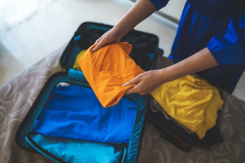 Eine Frau packt Kleidungsst&uuml;cke in einen kleinen Koffer, der auf einem Bett liegt.