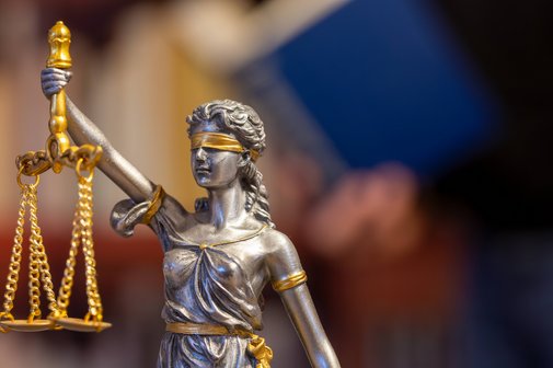 Nahaufnahme einer Justitia, im Hintergrund ließt ein Mann in einem Buch