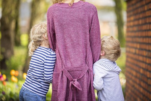Rückenansicht einer Mutter, die rechts und links ein kleines Kind an den Händen hält, die Kinder schmiegen sich an sie.