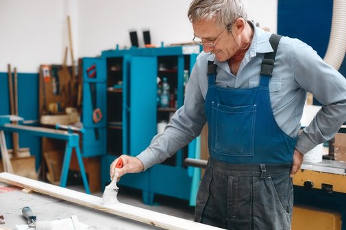Ein &auml;lterer Handwerker mit grauem Haar und Brille streicht eine Leiste mit wei&szlig;er Farbe, hat eine Hand in den R&uuml;cken gest&uuml;tzt, tr&auml;gt einen Blaumann
