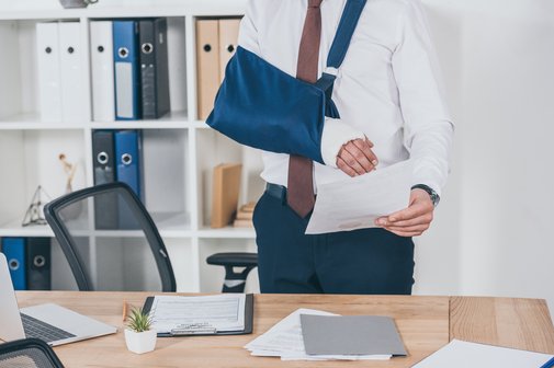 Krank am Arbeitsplatz Ein Geschäftsmann an seinem Schreibtisch, seine rechte Schulter ist in einer Schlinge