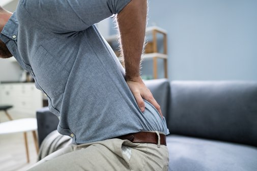 Rückenschmerzen Ein Mann steht vom Sofa auf, hält sich mit einer Hand den unteren Rücken als Symbol für Rückenschmerzen