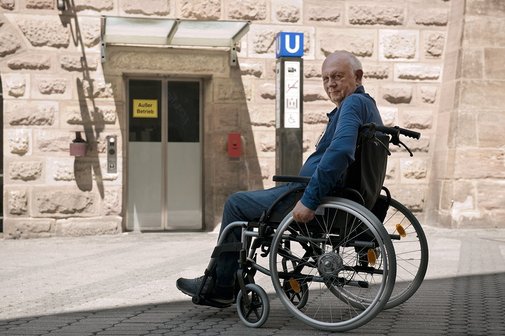 Ein Senior im Rollstuhl steht am unzugänglichen Eingang einer U-Bahn-Station. Dort ist ein Aufzug mit dem Hinweis "Außer Betrieb". 