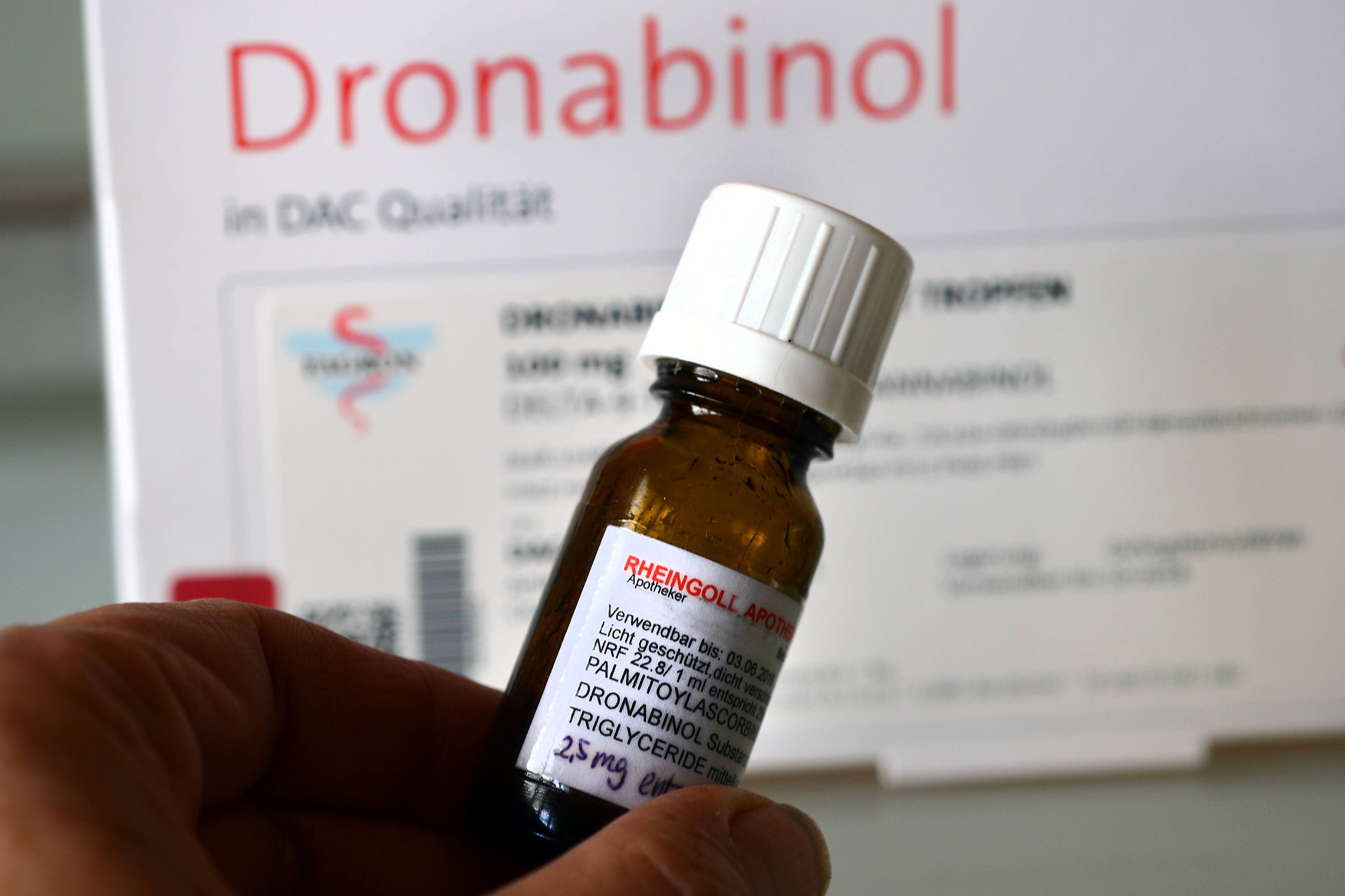 Eine Medikamentenpackung mit der Aufschrift "Dronabonil", im Vordergrund hält jemand ein Arzneifläschchen in der Hand.