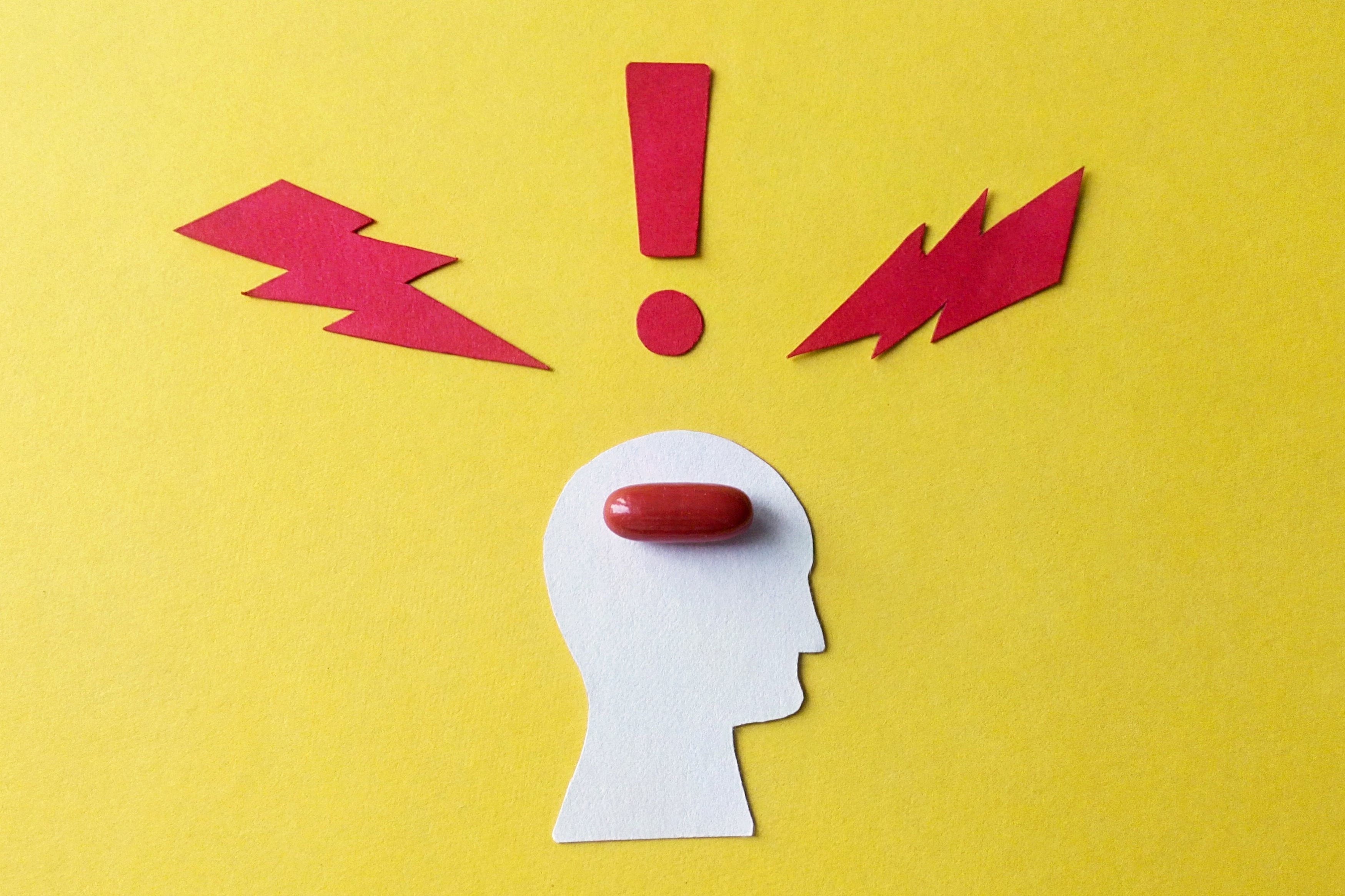 Symbol für Migräne und Kopfschmerzen: Ein aus Pappe ausgeschnittener stilisierter Kopf auf gelben Hintergrund, darin liegt eine rote Pille. Um den Kopf herum symbolisieren zwei rote Blitze und ein rotes Ausrufezeichen Schmerzen.