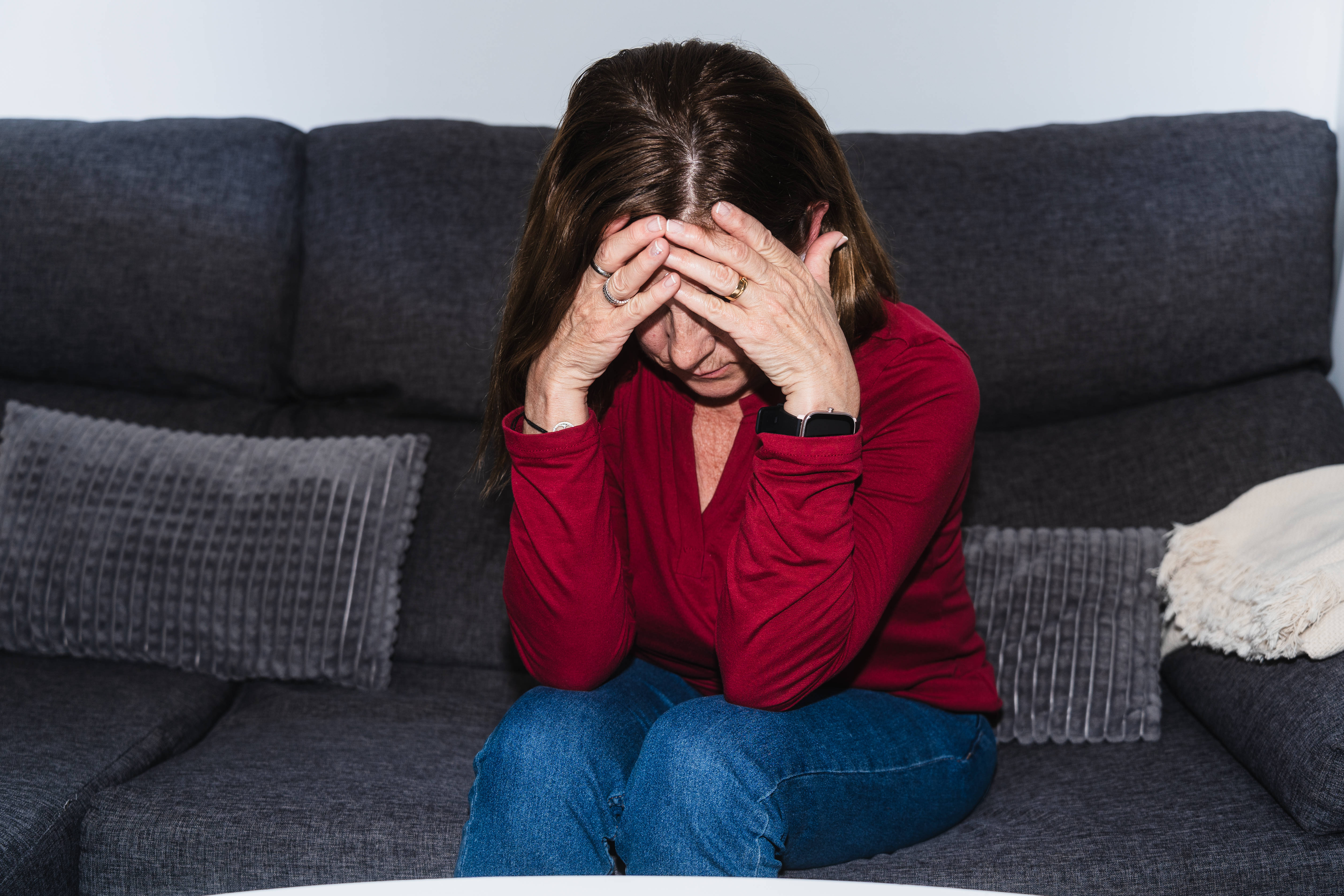 Eine ältere Frau sitzt auf einem Sofa, hat den Kopf gesenkt und hält sich die Hände vor das Gesicht. Sie sieht erschöpft oder traurig aus. 