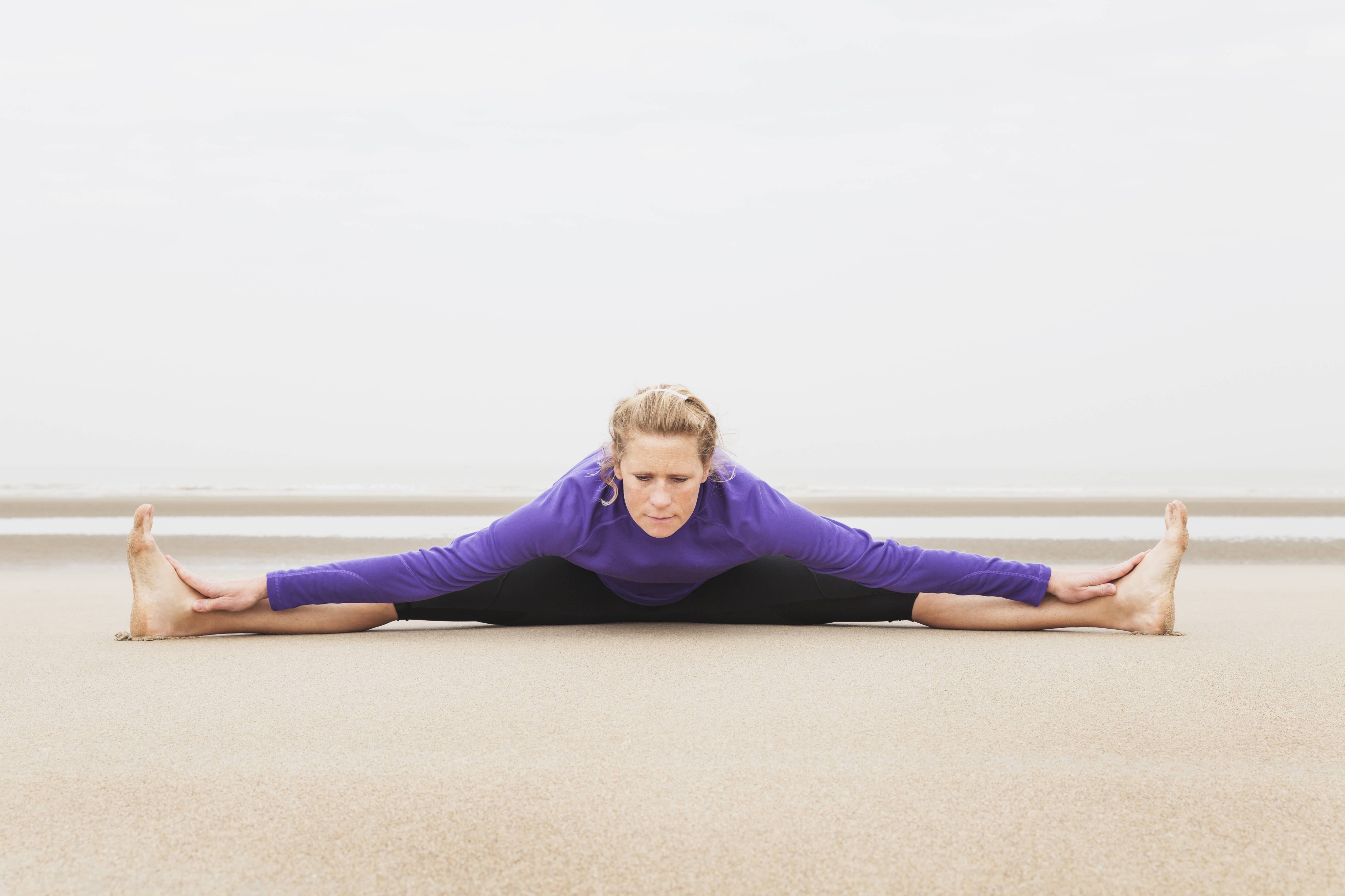 Eine Frau macht einen Spagat am Strand. Ihr gesicht sieht angestrengt und konzentriert aus. Ihre H&auml;nde ber&uuml;hren ihre Fu&szlig;r&uuml;cken, sie beugt sich weit nach vorne.