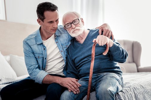 Wir setzen uns für eine menschenwürdige Pflege ein Sohn sitzt mit seinem älteren pflegebedürftigen Vater auf der Bettkante und hält ihn in den Armen