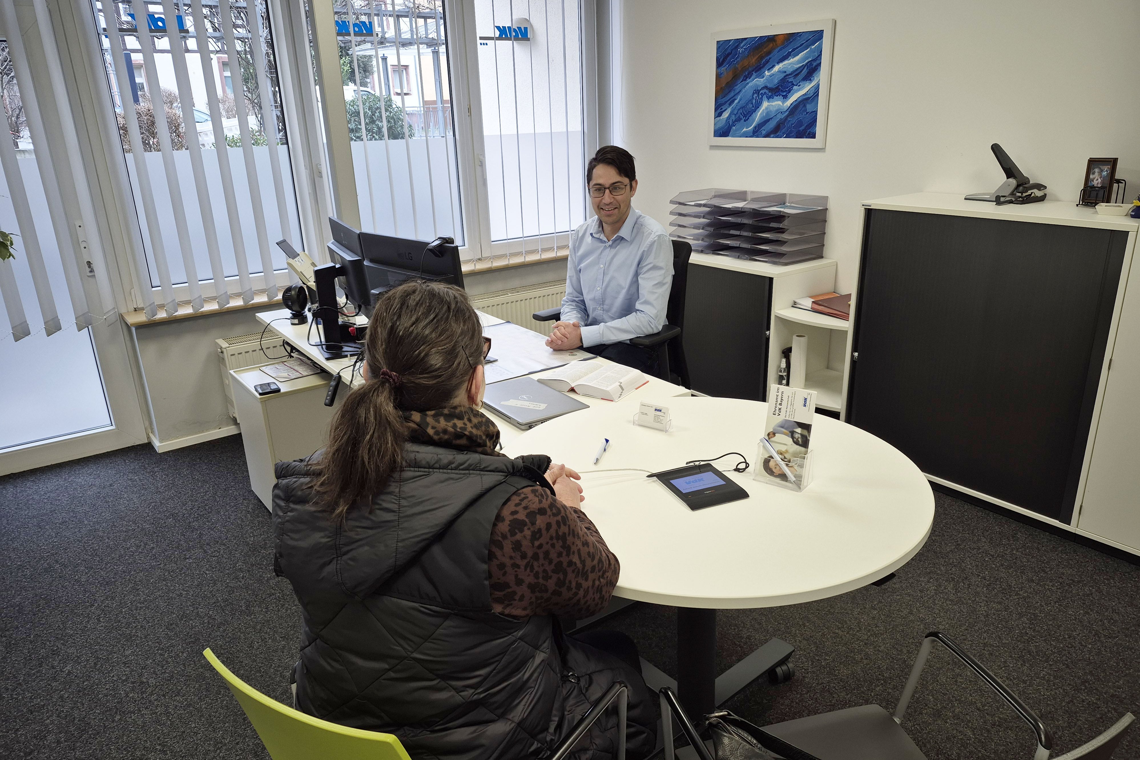In einem B&uuml;ro einer VdK-Kreisgesch&auml;ftsstelle l&auml;sst sich ein VdK-Mitglied beraten, das mit dem R&uuml;cken zum Betrachter an einem Schreibtisch sitzt. Der Frau gegen&uuml;ber sitzt ein VdK-Sozialrechtsberater, der sie aufmerksam und freundlich anblickt. 