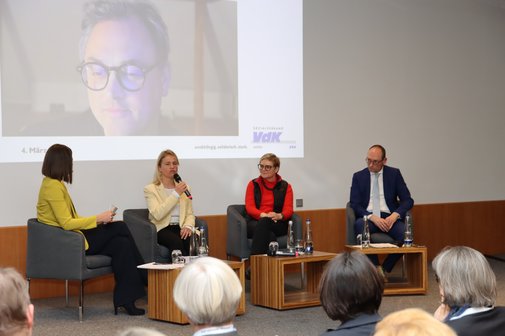 Auf dem Foto sieht man (von links): Moderatorin Isabella Kroth, VdK-Landesvorsitzende Verena Bentele, Parlamentarische Staatssekretärin im Bundesgesundheitsministerium Sabine Dittmar, Vorsitzender des Gesundheitsausschusses im Bayerischen Landtag Bernhard Seidenath und im Hintergrund auf einer Leinwand online zugeschaltet Mitglied der Regierungskommission Kliniken Prof. Dr. Christian Karagiannidis.
